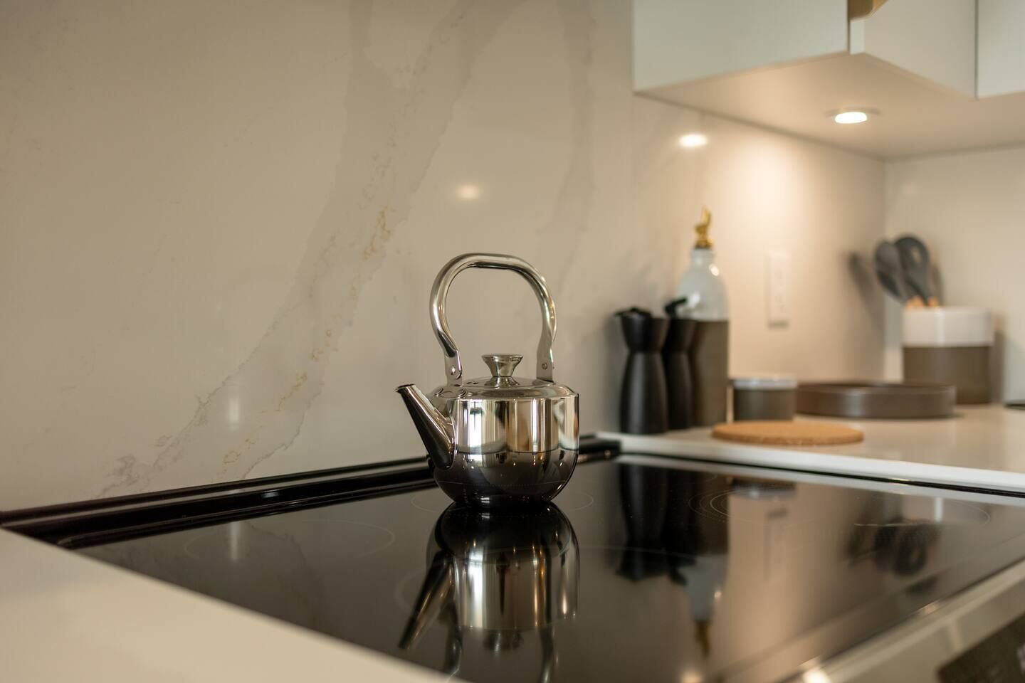A stainless steel kettle on a black stovetop in a modern kitchen with marble walls and contemporary appliances.