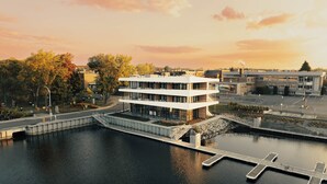 A modern multi-story building with a dock extending into a calm body of water, surrounded by trees and a parking area. - La Capitainerie - Marina - 304 - Bord de l'eau (Lac-Mégantic)