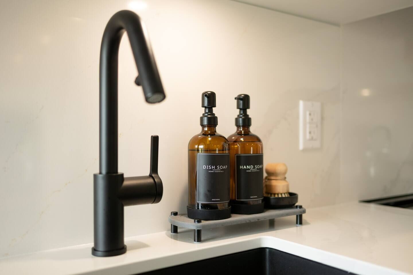 Eine moderne Küche mit einem eleganten schwarzen Wasserhahn, zwei Flaschen Geschirrspülmittel und Handseife sowie einer Holzbürste auf einer Arbeitsplatte.