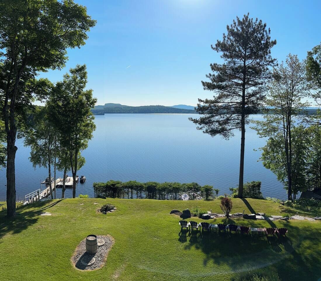 Vista a un lago con una zona cubierta de césped, una parrilla y una hilera de sillas Adirondack.
