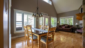 A dining area with a wooden table and chairs, a chandelier, and large windows with a view of trees. - Aux mille et une étoiles (Piopolis)