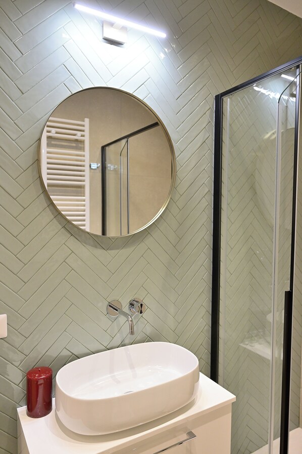 Une salle de bain avec un mur orné d’un motif en chevrons, un miroir rond, un lavabo blanc et une cabine de douche en verre.