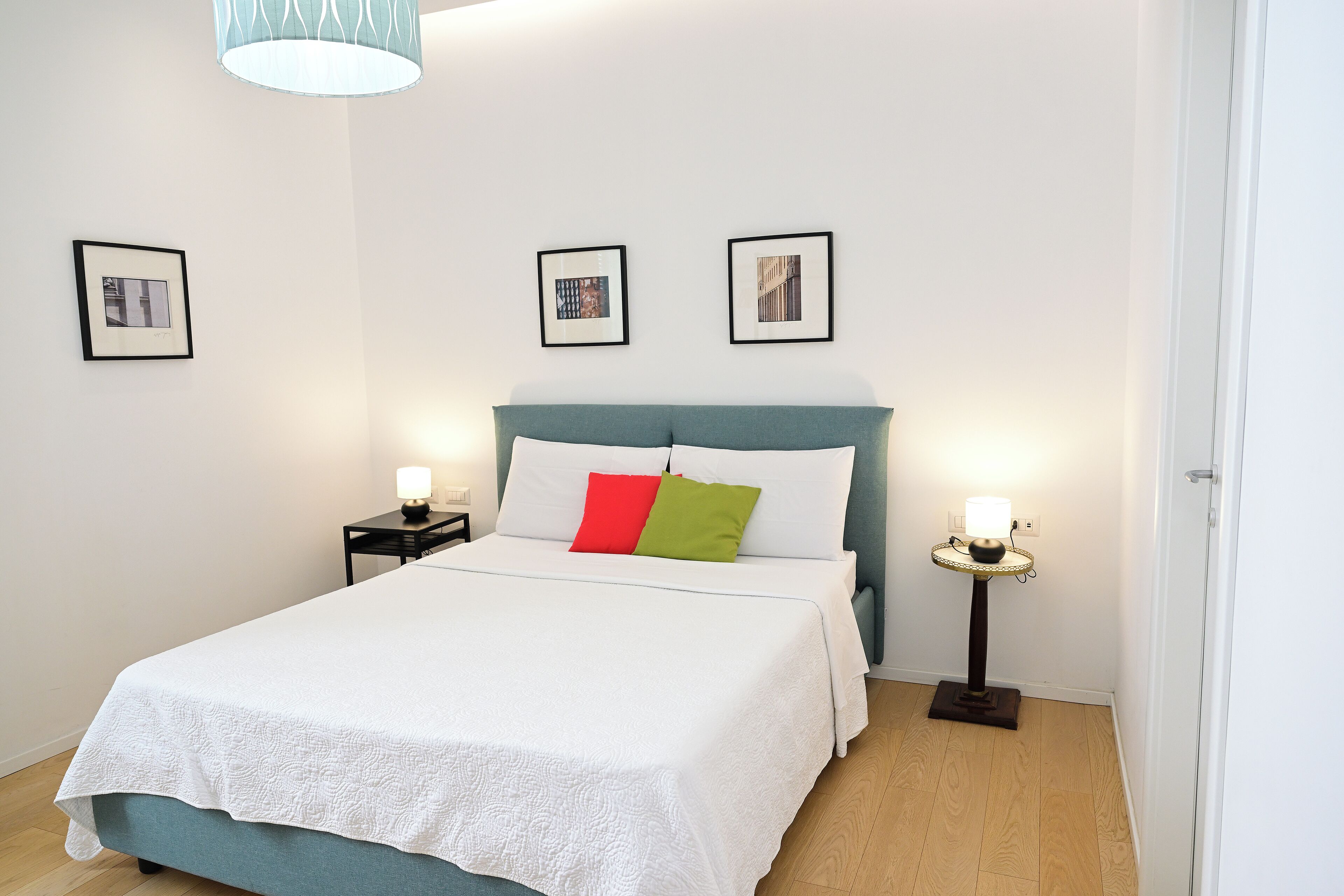 A neatly made bed with a white duvet and two pillows, a bedside table with a lamp, and two framed pictures on the wall.