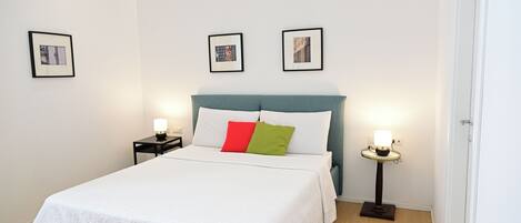 A neatly made bed with a white duvet and two pillows, a bedside table with a lamp, and two framed pictures on the wall.