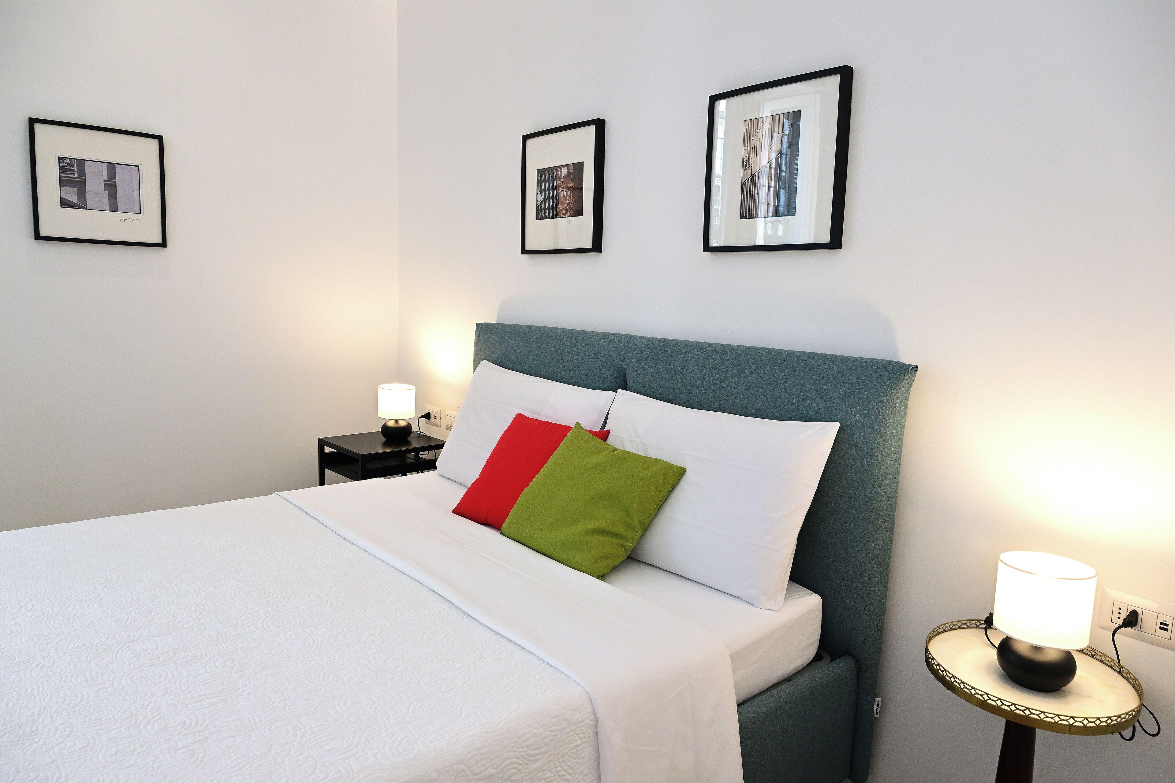 Un lit bien fait, avec une literie blanche, une tête de lit turquoise et un coussin rouge et vert. On peut voir des tableaux encadrés au mur, une lampe de chevet et une petite table avec une lampe.
