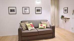 A living room with a grey sofa, three framed pictures on the wall, a light fixture, and a plant on a shelf.