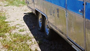 A vintage camper van parked on a dirt path with a lake and mountains in the background.
