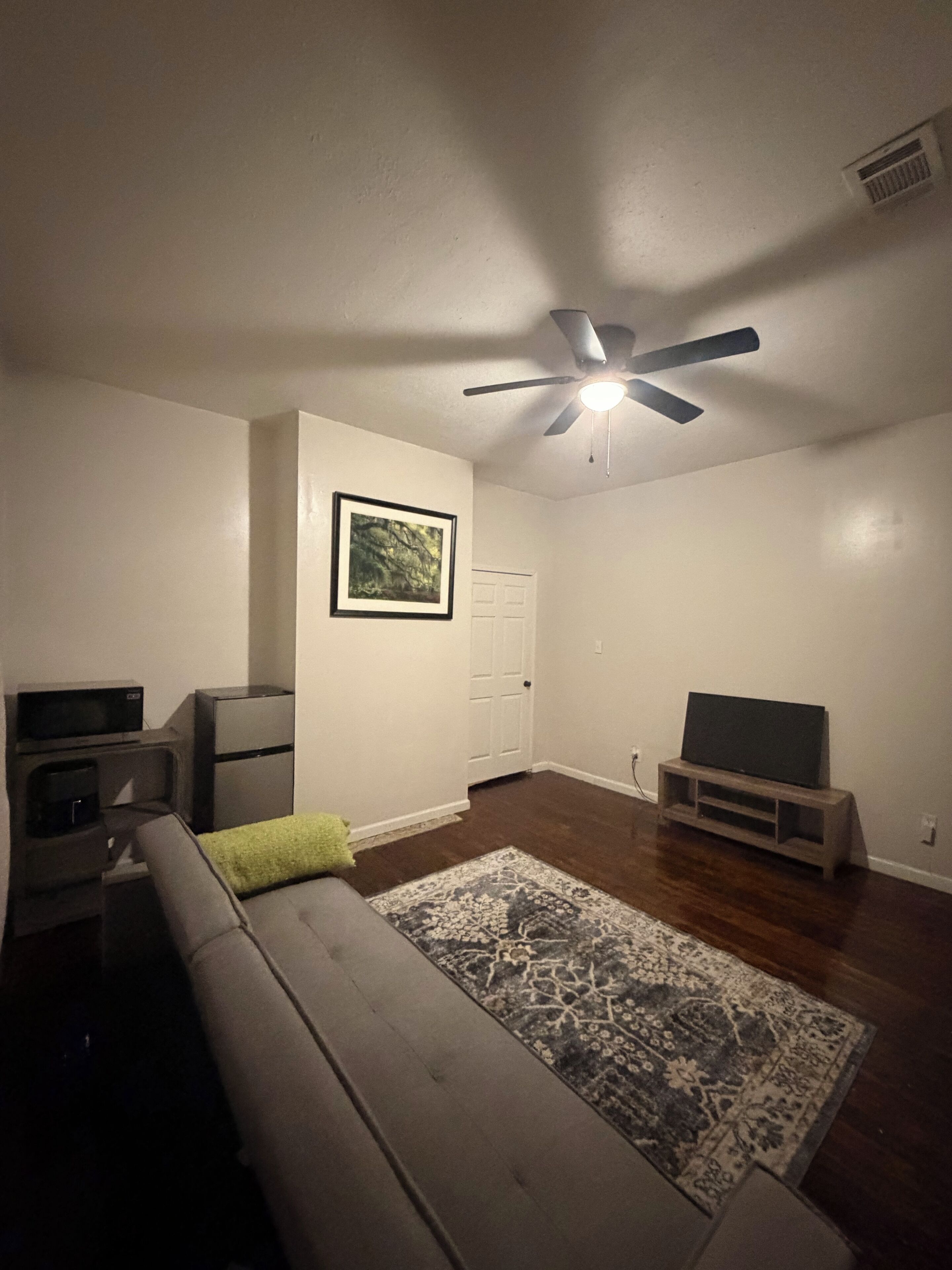 Una sala de estar con ventilador de techo, televisor sobre un mueble bajo, sofá, un archivador pequeño y un cuadro en la pared.