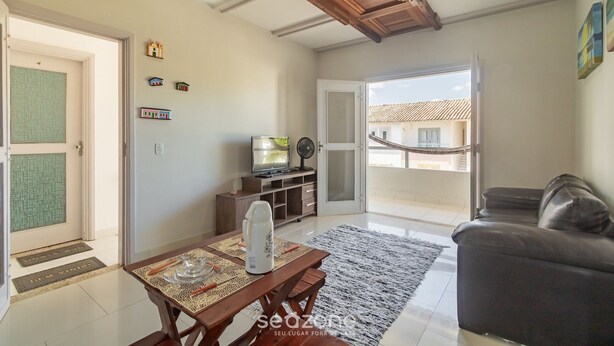 A living room with a wooden ceiling, a sofa, a TV on a stand, a dining table with a placemat, and a view of a balcony through a glass door. - Apt W/pool and Barbecue Near the Beach BA Art003 (Porto Seguro)