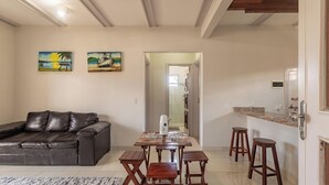 A living room with a leather sofa, a dining table with stools, and two paintings on the wall. - Apt W/pool and Barbecue Near the Beach BA Art003 (Porto Seguro)