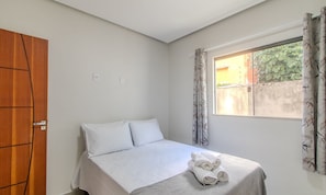 A single bed with white linens and a rolled towel, a wooden wardrobe, and a window with patterned curtains. - Apartamento Moderno a 280m da Praia AST0103 (Porto Seguro)