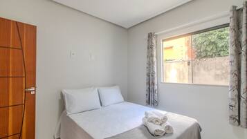 Uma cama de casal com roupa de cama branca, um guarda-roupa de madeira e uma janela com cortinas estampadas.