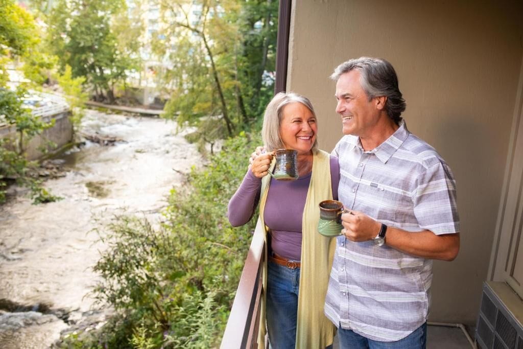 Un hombre y una mujer de pie junto a una ventana con vistas a un arroyo pedregoso y vegetación.