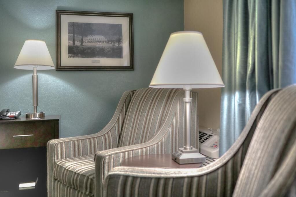 A hotel room with a striped armchair, a bedside table with a lamp, a framed picture on the wall, and a telephone on the table.