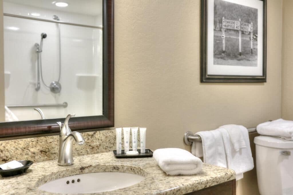 A bathroom with a granite countertop sink, a mirror, a framed black and white photo, a towel rack, and a shower area.