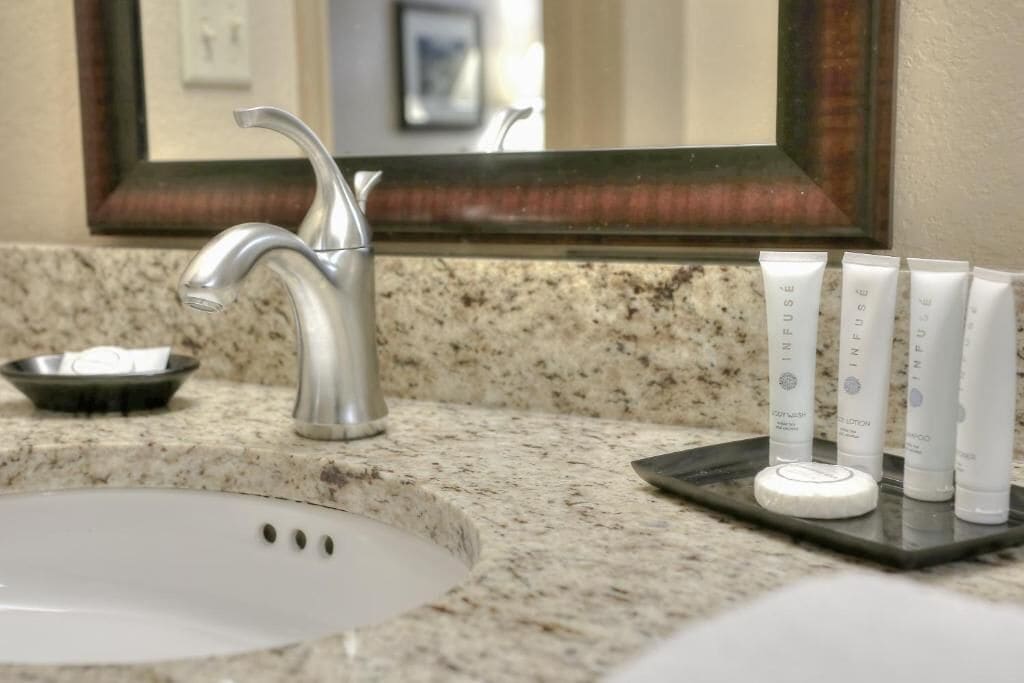 A bathroom sink with a granite countertop, a silver faucet, and a mirror above it. There are three tubes of shampoo and a soap dish on the counter.