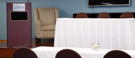 Een vergaderzaal met een tafel gedekt voor een vergadering, een spreekgestoelte, een gestreepte stoel en een televisie aan de muur.