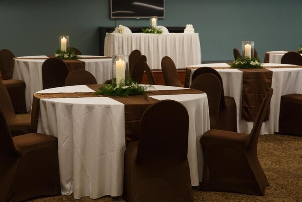 Une salle avec des tables rondes recouvertes de nappes blanches, des chaises marron et des bougies.