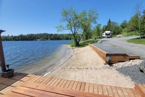 Una terraza junto al lago con un paseo de madera, una pequeña plataforma de madera y un área de grava que conduce a una casa.