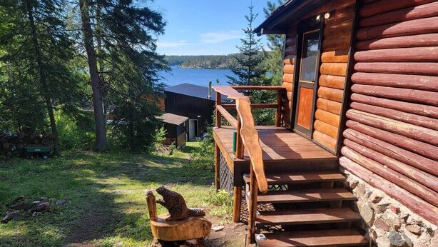 En timmerstuga med en trätrappa som leder upp till en veranda, omgiven av träd och med utsikt över en sjö.
