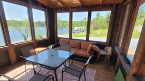 Eine Holzhütte mit einer gläsernen Veranda, einem Tisch, Stühlen und einer Couch mit Kissen.