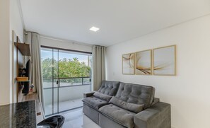 A modern living room with a grey sofa, a glass door leading to a balcony, and a minimalist decor.