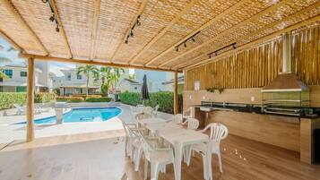 An outdoor patio with a pool, white plastic chairs, a wooden table, and a grill area.