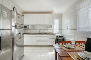 A modern kitchen with white cabinets, stainless steel appliances, and a dining area with wooden chairs and a table. - Spacious House w/ Pool in Guarajuba Cag0003 (Camaçari)