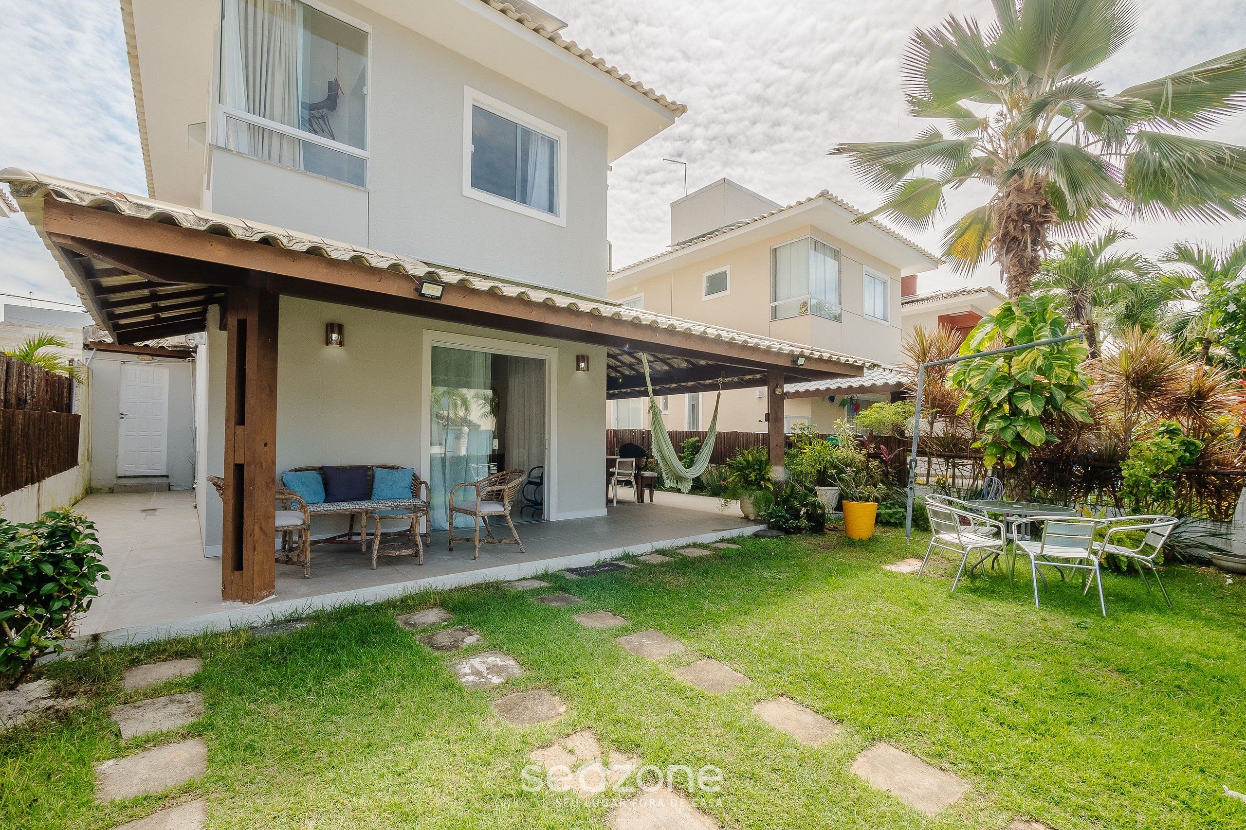 Una casa de dos pisos con un porche cubierto, mobiliario exterior y un jardín cuidado con palmeras y plantas en macetas.