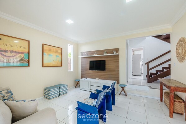 A living room with a sofa, a television mounted on a wooden wall, and a staircase leading to another level. - Spacious House w/ Pool in Guarajuba Cag0003 (Camaçari)