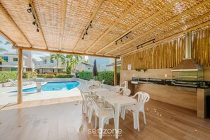 Un patio exterior con piscina, sillas de plástico blanco, una mesa de madera y zona de barbacoa.