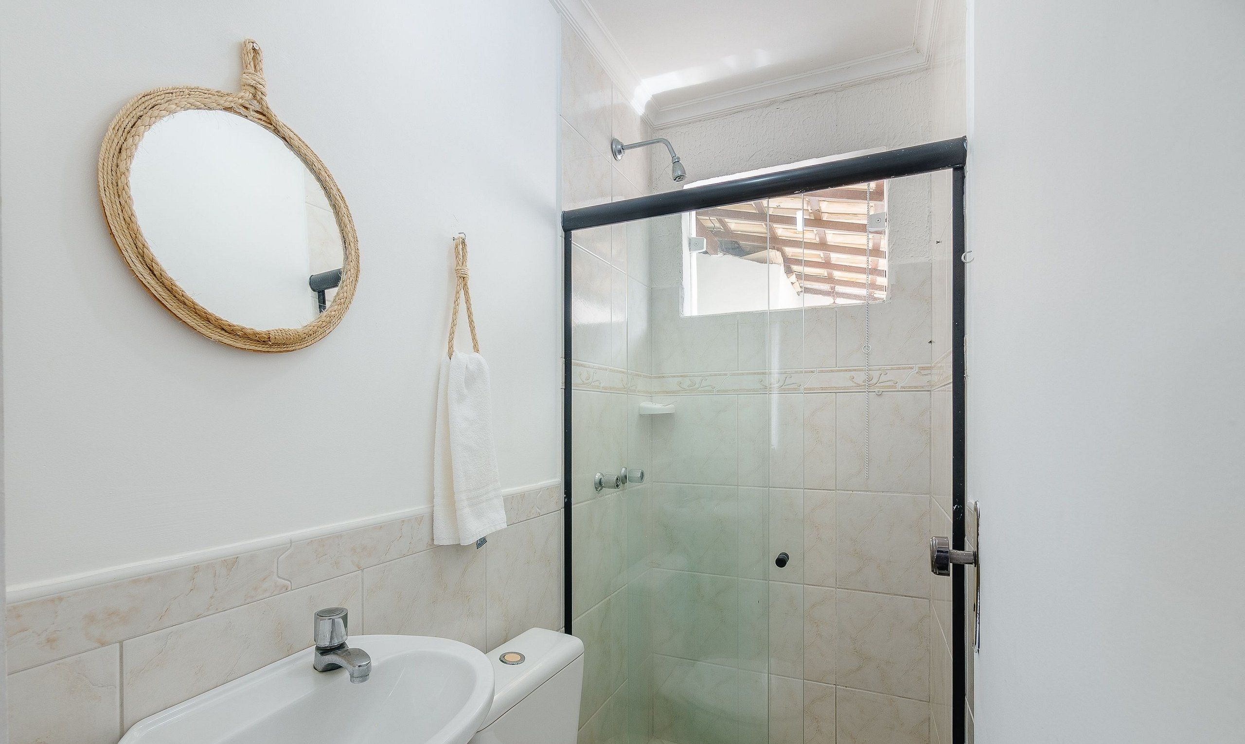Un baño con ducha de vidrio, un lavamanos blanco y un espejo redondo con marco de cuerda.