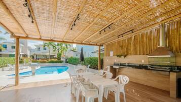 Un patio exterior con piscina, sillas de plástico blanco, una mesa de madera y zona de barbacoa.