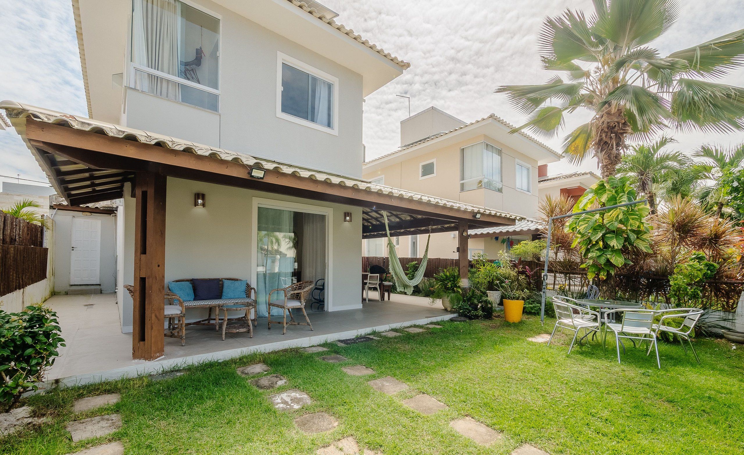 A two-story house with a covered patio, outdoor seating, and a well-maintained garden with palm trees and potted plants.