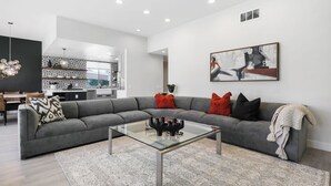 A modern living room with a grey sectional sofa, a glass coffee table, and a kitchen in the background. - GATED, Spacious 2-bedroom condo in Indian Wells close to restaurants and more. (Indian Wells)