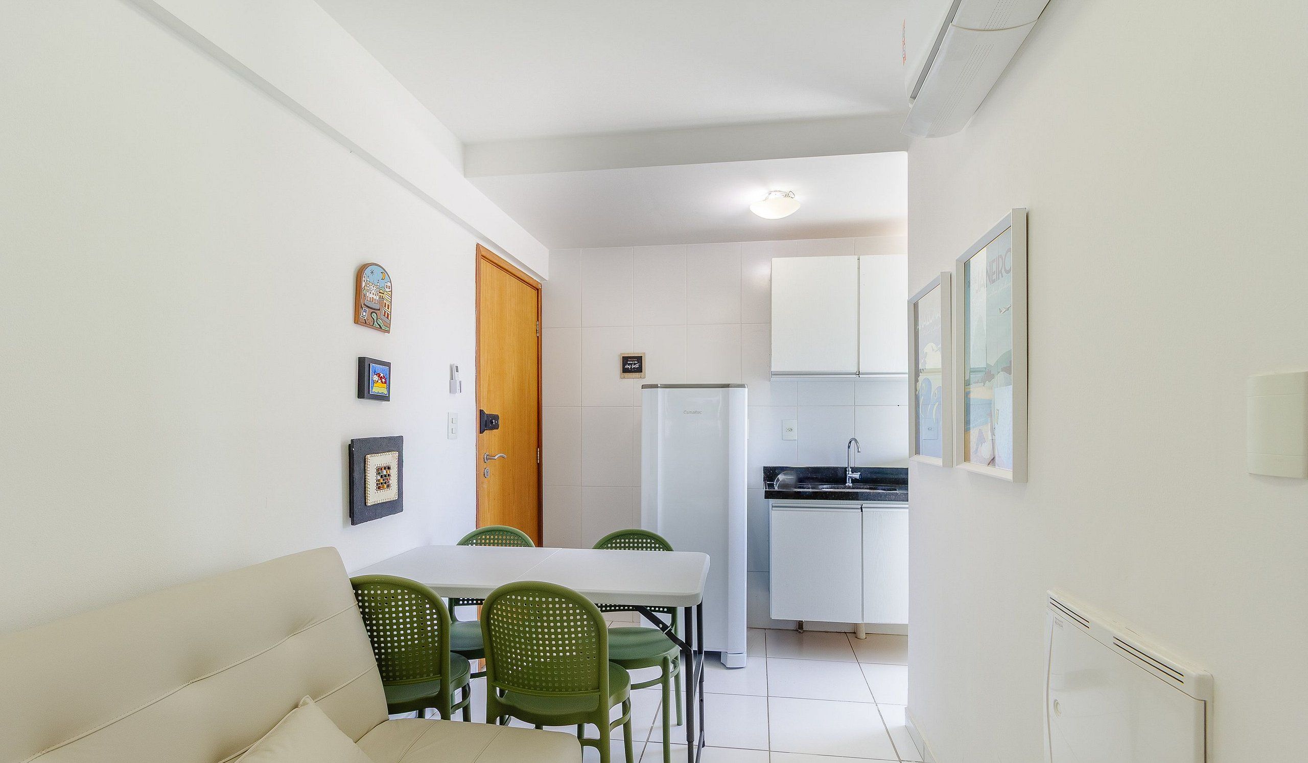 A compact kitchen with white cabinets, a refrigerator, and a sink. There is a dining table with four green chairs and a beige sofa in the foreground.