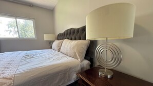 Ein ordentlich bezogenes Bett mit weißer Decke und Kissen, ein gepolstertes Kopfstück, ein Nachttisch mit moderner Lampe und ein Fenster mit Blick auf Bäume.