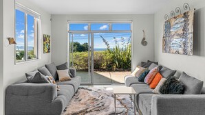 Una sala de estar moderna con sofás grises, una mesa de centro y un gran ventanal que da a un balcón con plantas y vistas al mar.
