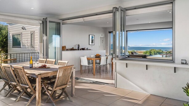 Un área de comedor exterior moderna con vistas al mar, una cocina con puertas de vidrio correderas y un patio con mesa y sillas.