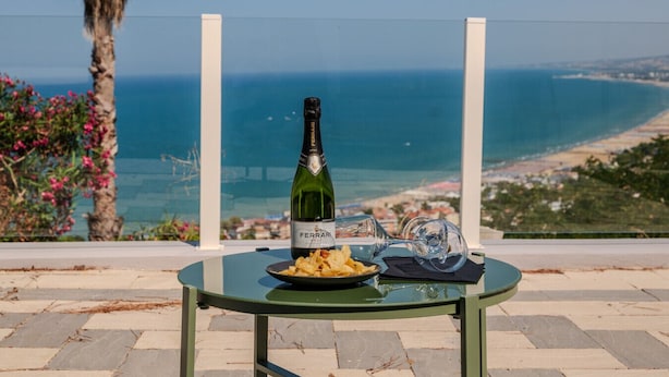 Ein Tisch mit einer Weinflasche, einem Glas und einem Teller Essen auf einer Terrasse mit Blick auf einen Strand und das Meer.
