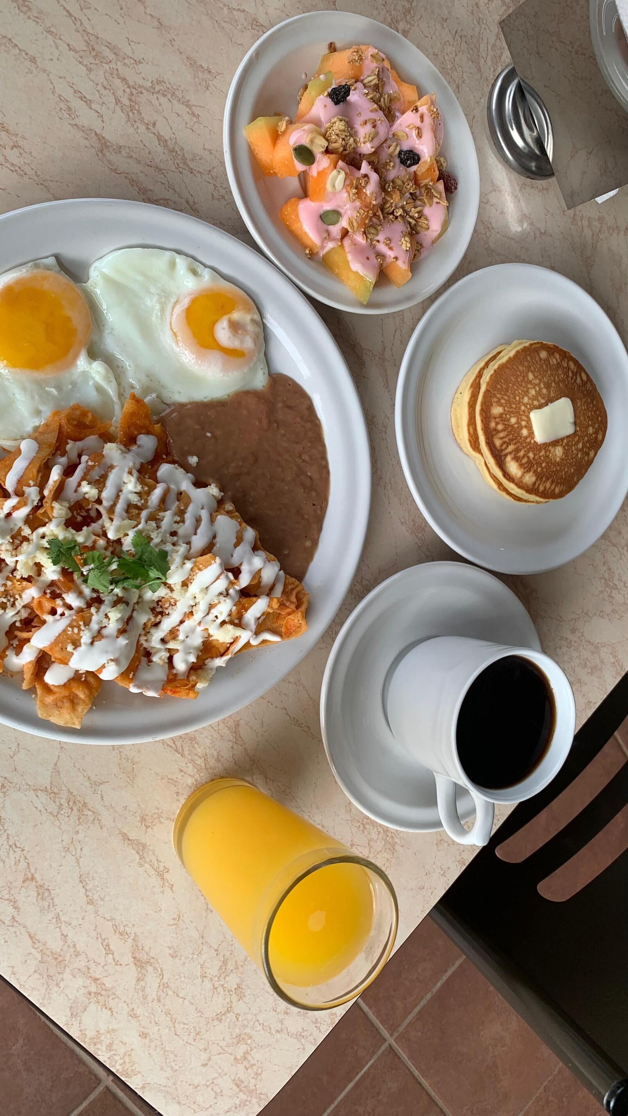 Una mesa de desayuno con platos de huevos, chilaquiles, panqueques y un vaso de jugo de naranja.