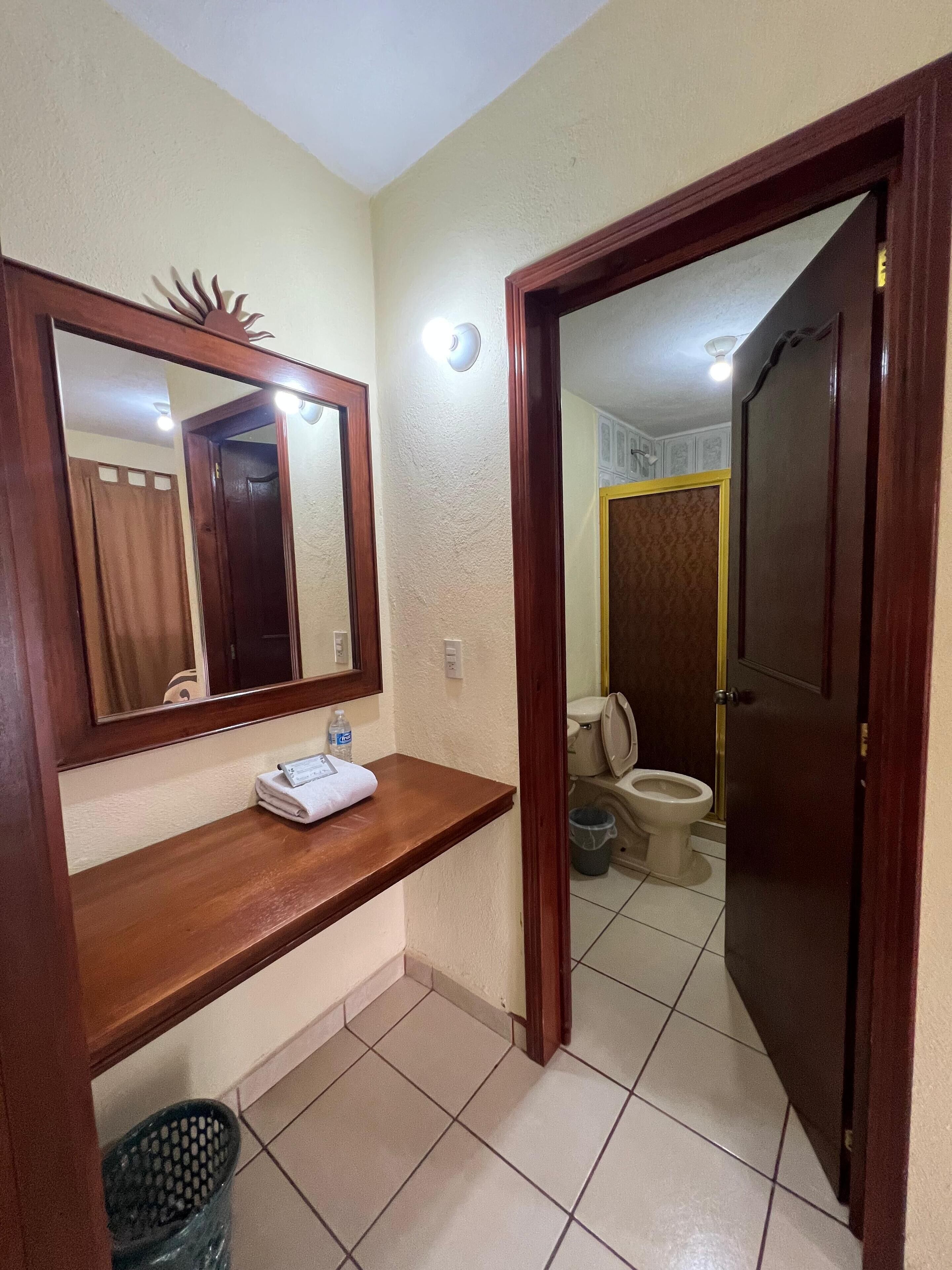 A bathroom with a wooden vanity, a mirror, a towel, a trash bin, and a toilet visible through an open door.