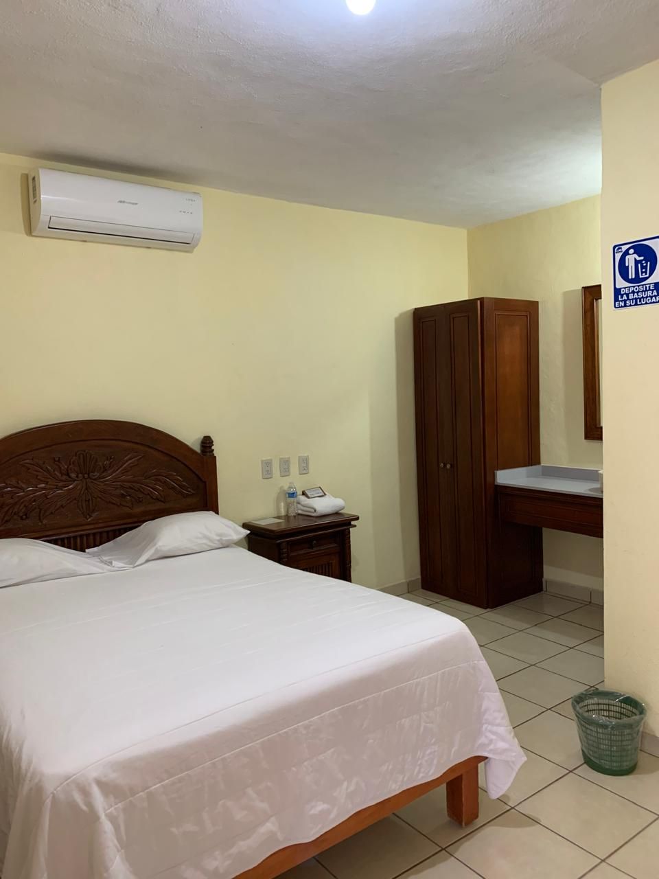 Habitación de hotel con una cama, un armario de madera, un escritorio con espejo y un cesto de basura.