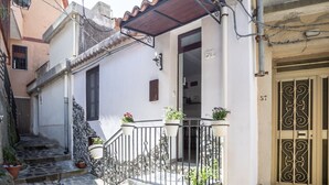 A narrow street with a white building featuring a small balcony with potted plants, a metal gate, and a stone staircase. - House in the mountains near the sea (Itala)