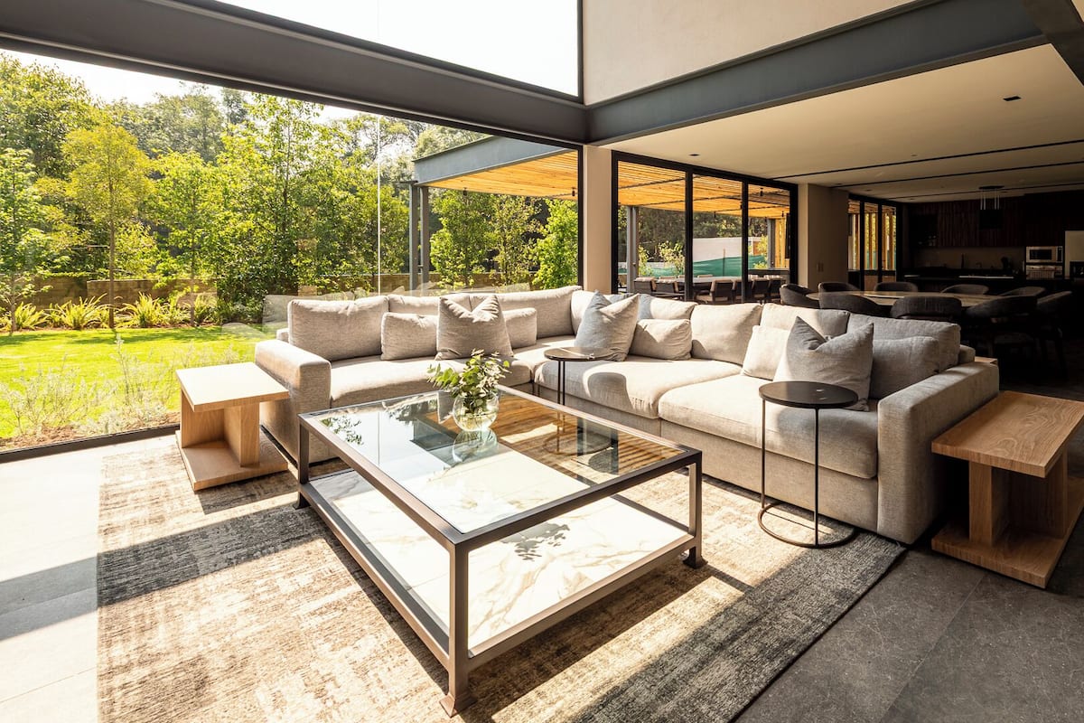 A modern living room with a large sofa, a glass coffee table, and a view of a garden through large windows.
