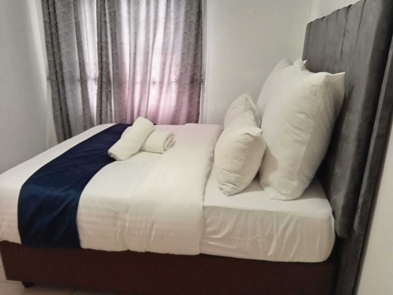 A neatly made bed with white bedding and a dark blue accent, a wooden headboard, and a window with curtains.