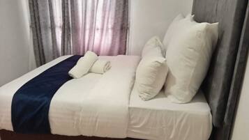 A neatly made bed with white bedding and a dark blue accent, a wooden headboard, and a window with curtains.