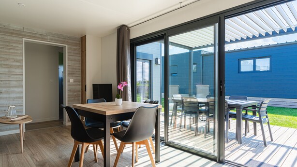 Ein moderner Essbereich mit einem Holztisch und Stühlen, einer Kübelpflanze und Blick auf ein blaues Haus durch große Schiebeglastüren.