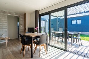 A modern dining area with a wooden table and chairs, a potted plant, and a view of a blue house through large sliding glass doors.