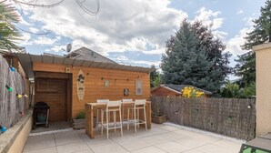 A wooden house with a tiled patio, a wooden fence, and a large tree in the background.